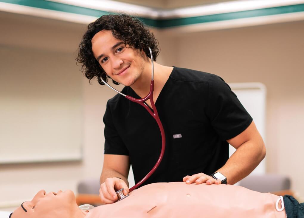 Elias Sanchez leans over a medical human manikin holding the stethoscope to it's chest